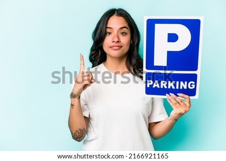 Young hispanic woman holding parking placard isolated on blue background showing number one with finger.