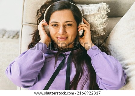 A woman in headphones listens to her favorite music at home on the couch. Listening to meditations, e-books and podcasts in your spare time. The woman lies on the couch and smiles. View from above