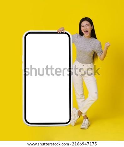 Full body asian girl stand near huge, giant smartphone with white screen happy making yeah, winner hand gesture dressed in casual isolated on yellow background. Free space mock up. 