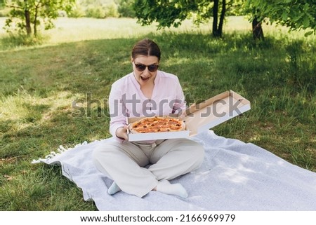 Happy young woman holding hot pizza in box on nature background. Attractive girl feels hungry and loves the taste. Entire pizza for myself. Fast food banner