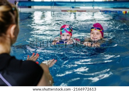 Children learn to swim with board in pool under guidance of coach. Swimming lesson. Active kids are playing in water. Girls in goggles and swimming cap.