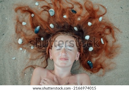 Red-haired girl resting on a sunny sea sandy beach. Young woman standing by the sea and lying on the sea sand with shells.