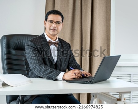 A businessman looks at the camera with a smile. The man works in the office for a laptop. A business person looks welcome. The man works at the table.