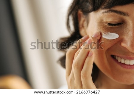 Close-up happy young Caucasian woman smiling with her eyes closed applying cream on her face. Brunette moisturizes her skin with cosmetics. Home spa care concept