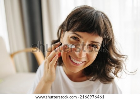 Close-up of beautiful young caucasian girl looking at camera applying cream on her face indoors. Brunette in white t-shirt performs morning ritual. Wellness and self care concept Royalty-Free Stock Photo #2166976461