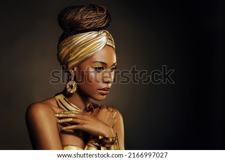Portrait Closeup Beauty fantasy african woman, face in gold paint. Golden shiny black skin. Fashion model girl mixed race. Glamorous arab turban, jewellery accessories. Professional metallic makeup. Royalty-Free Stock Photo #2166997027