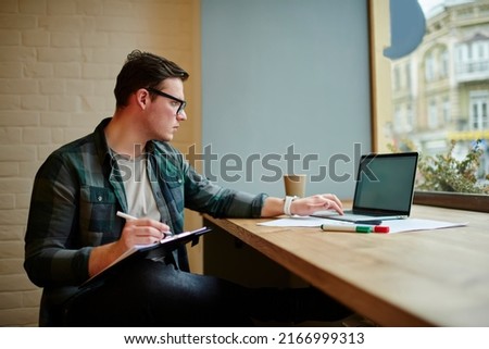 Young smart freelancer in glasses working with documents and laptop computer at desk. Male student learning online with netbook Royalty-Free Stock Photo #2166999313