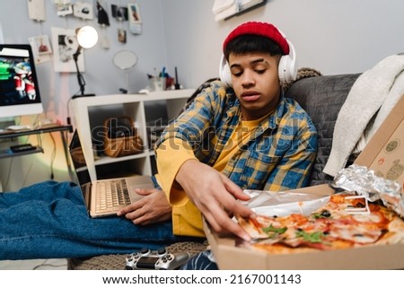 Middle-eastern teenage boy eating pizza and using laptop while sitting on sofa at home