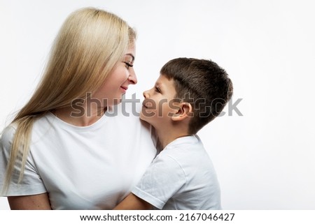 Mom and little son hug and smile. A beautiful blonde and a boy of 7 years old in white T-shirts looks at each over. Love and tenderness. Light background.