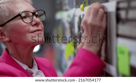 Close up of mature editor pin pictures to magnetic board in fashion magazine headquarters. Businesswoman work with mood board