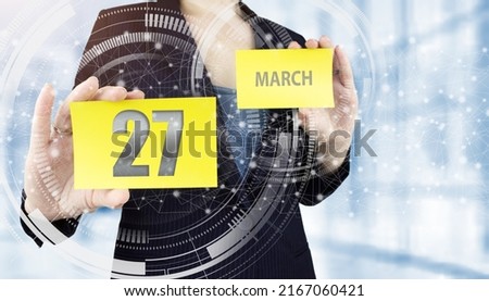 March 27th. Day 27 of month, Calendar date. Business woman hand hold yellow sheet with calendar date on blurred office background.  Spring month, day of the year concept