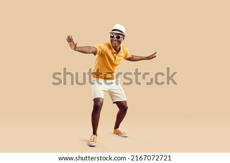 Cheerful stylish african american man having fun dancing on light beige background. Funny smiling dark skinned man in sneakers, shorts, polo shirt and hat with sunglasses. Full length. Banner. Royalty-Free Stock Photo #2167072721