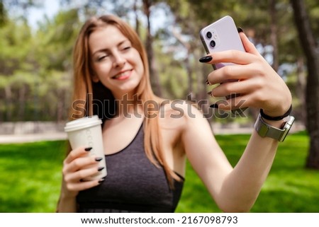 Cute brunette woman wearing sports bra standing on city park, outdoors taking selfie and holding takeaway coffee mug. Self portrait for social media. Outdoor sport concepts.