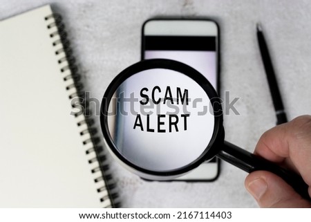 A man's hand holds a black magnifying glass by the handle on a smartphone background with scam alert text.