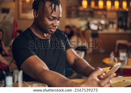 African american man in casual outfit using a smartphone and reading credit card number while sitting at cafe table. Online payment and shopping. Ordering food online, booking tickets.