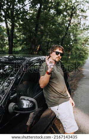 Stylish long-haired hippie man in sunglasses stands near a black car on the road in nature. Photography, travel, business, portrait.