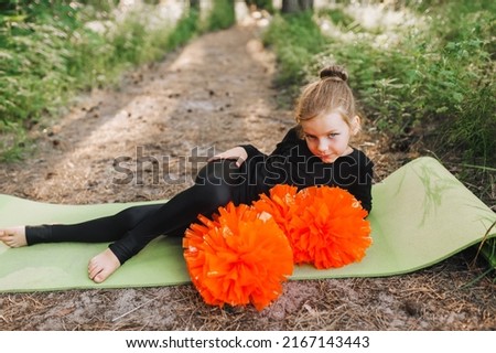 A tired, smiling girl, a child cheerleader in a black suit lies, resting on a green rug in the forest with large orange pom-poms in her hands. Sports training for cheerleading. Photography, concept.