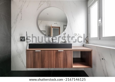 Modern design bathroom with wooden cabinets, black porcelain sink, circular mirror frame, chrome metal fixtures and fittings and black marble flooring Royalty-Free Stock Photo #2167146125