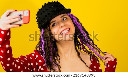 Young beautiful woman in red dress, black cap takes selfie on mobile phone. Vivid pretty female posing, photographing of themself on smartphone in studio on yellow background