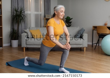 Positive sporty senior woman keeping fit at home, doing lunges, exercising on yoga mat in living room and smiling. Staying healthy at retirement Royalty-Free Stock Photo #2167150173