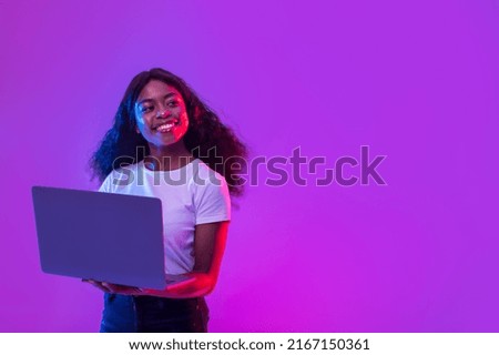 Portrait of cheerful millennial African American lady in casual wear using laptop computer for online work, education or communication, looking at empty space in neon light