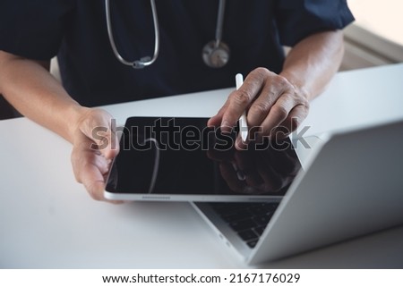 Doctor using digital tablet and working on laptop computer on table at doctor's office in hospital. Sugeon recording patient's information on tablet, electronic medical record system concept