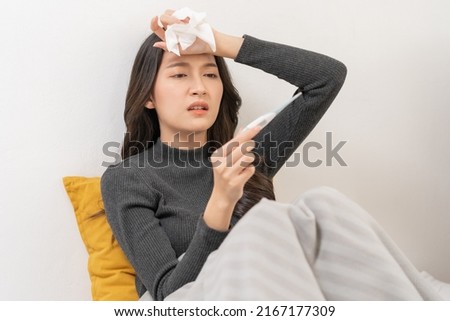 Sick, influenza asian young woman, girl headache have a fever, flu and check thermometer measure body temperature, feel illness sitting on sofa bed at home. Health care person on virus, covid-19. Royalty-Free Stock Photo #2167177309