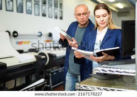 Female publishing facility worker reading operation manual for large printer. Her male co-worker standing beside. Royalty-Free Stock Photo #2167178227