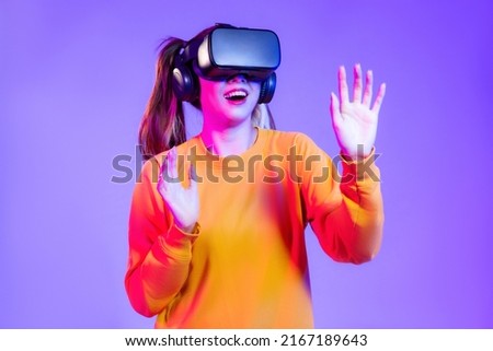 Young asian woman in orange sweatshirt wearing black vr headset watching playing and touching on the purple background. Metaverse concept vr future girl.