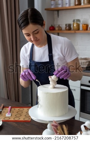 Woman in an apron aligns cream under chocolate bear on cake. Selective focus. Photos about confectioners, food, hobbies.