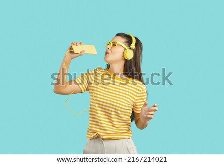 Happy carefree young woman in striped orange white T shirt and yellow headphones standing against turquoise background, holding mobile phone, listening to music, singing, dancing and having good time