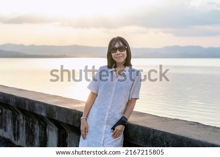 Woman traveler looking at the camera and standing on the street of the dam with a sunset background on a vacation trip. A female Asian tourist enjoying nature on holiday. Freedom concept
