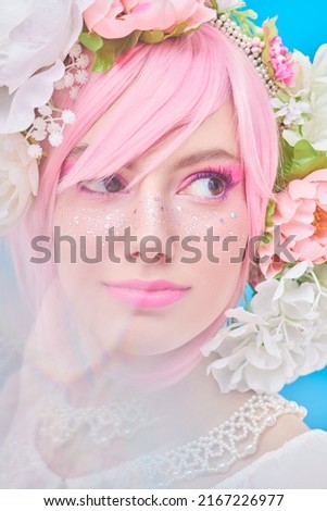 Beauty, makeup and hairstyle. Portrait of a pretty teen girl with bright pink make-up posing in colored pink wig and flower wreath on head. Studio portrait on a blue background.