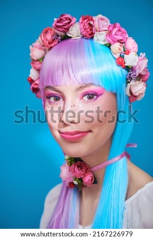 A pretty, smiling girl with bright pink make-up posing in colored violet-blue wig and flower wreath on head. Beauty, makeup and hairstyle. Blue background.