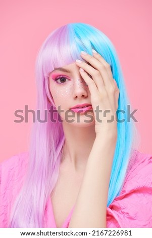 Hairstyling, hair dye. Portrait of a beautiful girl with colored purple-blue hair and bright pink makeup posing on a pink background covering her eye with her hand. Beauty, fashion. 