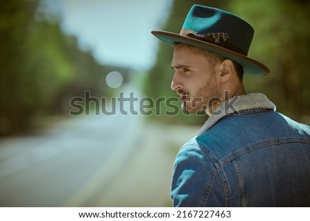 Portrait of a handsome bearded man in denim jacket and a hat standing by the highway. Denim fashion. Road adventures, hitchhiking, autotrip.  Royalty-Free Stock Photo #2167227463