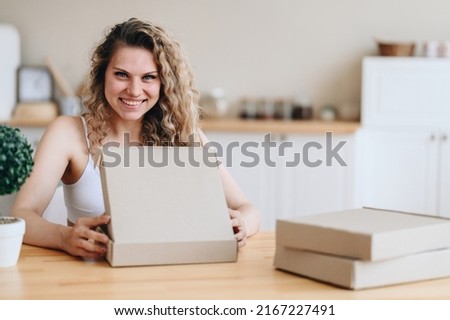 Happy smiling woman opens a delivery box. A woman sits at a wooden table in a bright home interior. Blurred kitchen background. Concept of Clearing parcels at home