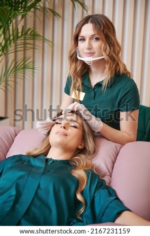 Portrait of beauty specialist comping client eyelashes after extension procedure. Female eyelashes technician in protective face mask combing woman lashes with disposable mascara brush in beauty salon Royalty-Free Stock Photo #2167231159