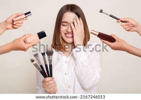Image of the hands of several beauticians holding their respective equipment giving makeover to beautiful smiling woman with charming smile, standing and covering eye with palm.