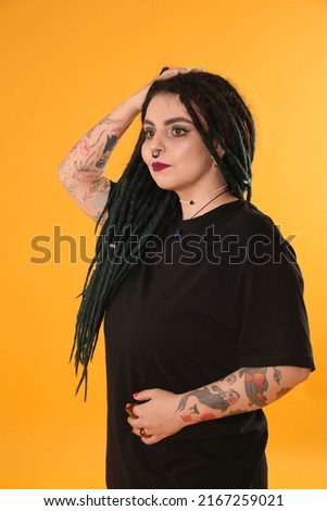 Beautiful young woman with tattoos on arms, nose piercing and dreadlocks against yellow background