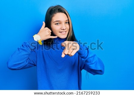 Young brunette girl wearing turtleneck sweater smiling doing talking on the telephone gesture and pointing to you. call me. 