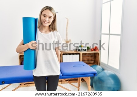 Young chinese woman smiling confident holding yoga mat at clinic