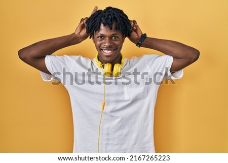 Young african man with dreadlocks standing over yellow background posing funny and crazy with fingers on head as bunny ears, smiling cheerful 