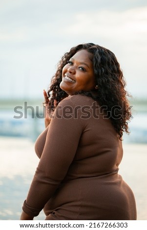 Vertical closeup portrait of curly smiling plump African American dark skin woman looking at camera, pose against blue sky. Interracial, mixed race female model. Ethnicity, diversity, body positive Royalty-Free Stock Photo #2167266303