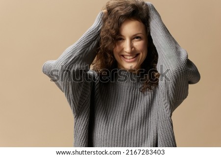 Cheerful smiling cute curly beautiful female in gray warm casual sweater touches head posing isolated on over beige pastel background. Fashion Sale offer. People emotions concept. Copy space