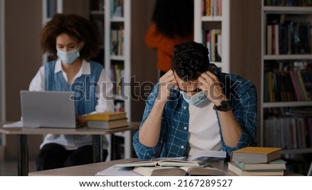 Tired overworked young arab male student in protective mask sits at desk in library does homework writes notes prepares for exam makes mistake grabs head with hands feels shock frustration failure