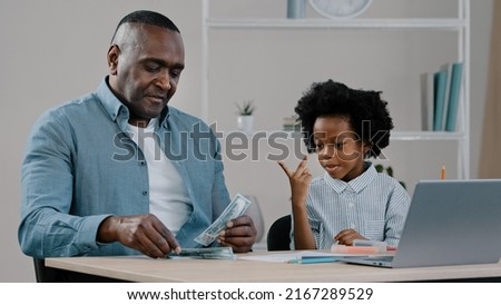 Cheerful adult father and little cute daughter sitting at home dad teaches kid girl to count shows dollars cash bills caring parent helps child learn how to save money basic math school preparation Royalty-Free Stock Photo #2167289529