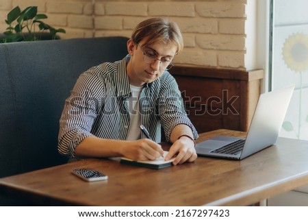 Smiling man with glasses sitting at table in cafe with laptop computer and writing something