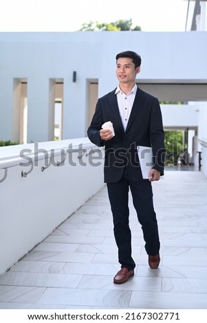 Full length portrait of elegant asian businessman holding take away coffee cup and walking outside modern office building