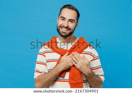 Young fun smiling satisfied cheerful kind-hearted man 20s in orange striped t-shirt looking camera put folded hands on heart isolated on plain blue background studio portrait. People lifestyle concept